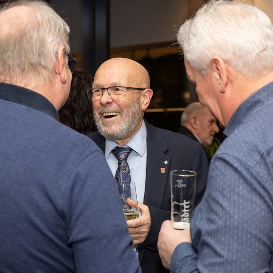 Drei Männer unterhalten sich. Der Mann in der Mitte hält ein Glas Bier. Er trägt eine Krawatte und eine Brille. Der Mann auf seiner linken und rechten Seite hält ebenfalls ein Glas Bier. Sie lächeln alle.