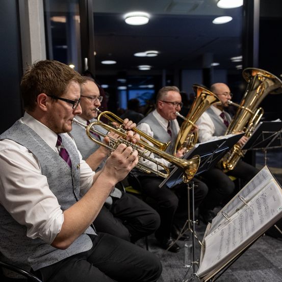 Eine Gruppe von Musikern, darunter ein Trompeter, tritt auf der Bühne auf. Sie sind in formeller Kleidung gekleidet und spielen von Notenblättern. Der Raum hat große Fenster und ein modernes Innendesign.