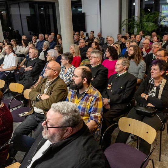 Eine große Menschenmenge sitzt in einem Raum, einige tragen Brillen, Uhren und Halsketten. Eine Frau vorne hält eine schwarze Tasche. Der Raum hat Stühle, eine Säule, eine Pflanze und eine Projektionswand.
