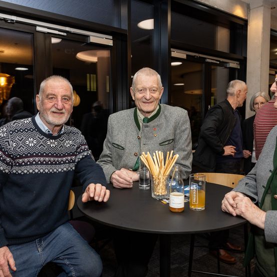 Zwei ältere Männer sitzen in einem Restaurant an einem Tisch, lächeln und schauen in die Kamera. Es gibt Gläser Bier und eine Flasche auf dem Tisch. Dahinter stehen mehrere Menschen und unterhalten sich.