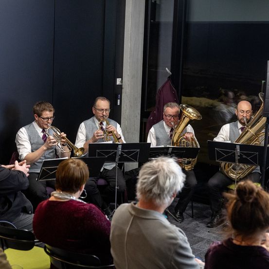 Eine Gruppe von Musikern, darunter Trompeter und Tubaspieler, tritt drinnen auf. Sie sitzen vor Notenständern und werden von einem Publikum beobachtet. Der Raum ist gut beleuchtet, und es gibt Reflexionen von Menschen draußen auf den Glasfenstern.