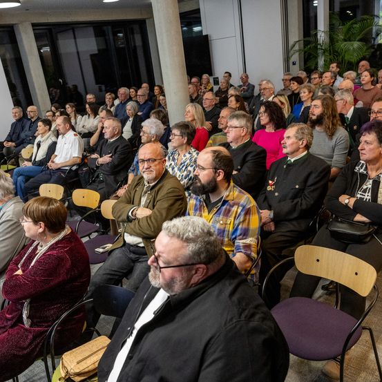 Eine große Menschenmenge sitzt in Stühlen in einem Innenraum, möglicherweise einem Auditorium. Viele tragen formelle Kleidung, Brillen und einige haben Gesichtsbehaarung. Eine Pflanze ist im Hintergrund sichtbar.