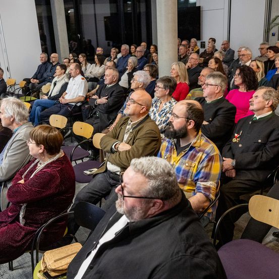 Ein überfüllter Raum mit Menschen in Reihen, die etwas beobachten. Viele mit Brille, einige mit Bärten. Die Stühle sind aus Holz, und der Raum hat weiße Wände.