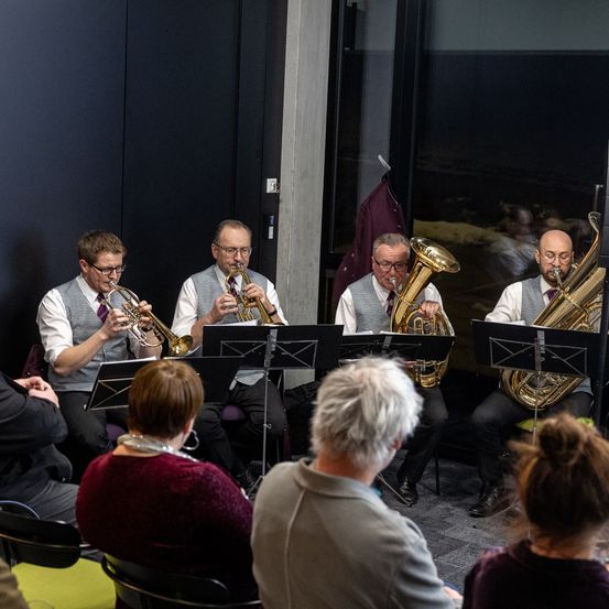 Eine Gruppe von Musikern, darunter Trompeten und Tubas, tritt vor einem Publikum auf. Sie sitzen vor Notenständern und scheinen in einer Probe zu sein.
