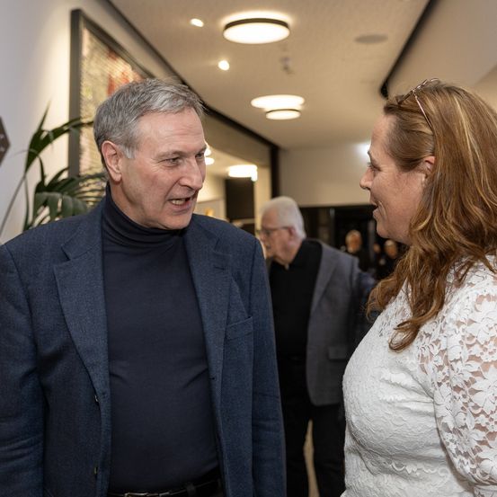 Ein älterer Mann in einem marineblauen Blazer und Rollkragenshirt spricht mit einer Frau in einem weißen Spitzenkleid, während andere Leute im Hintergrund durch einen Flur gehen.