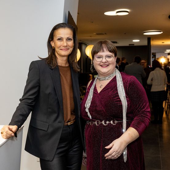 Zwei Frauen stehen nebeneinander. Eine Frau trägt einen schwarzen Blazer und ein braunes Hemd, die andere eine burgunderfarbene Robe mit einem silbernen Gürtel.