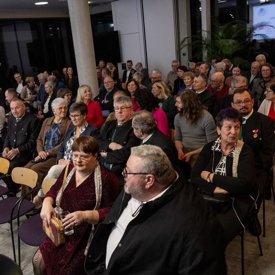 Eine große Zuhörerschaft sitzt in Reihen bei einer Veranstaltung und schaut auf eine Bühne mit einer Projektionsleinwand, während die Leute sich unterhalten und im Raum herumschauen.
