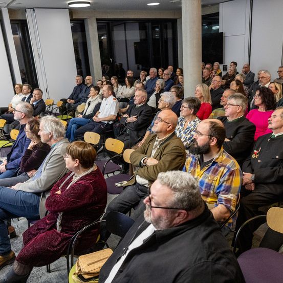 Ein großes Publikum sitzt in Reihen von Stühlen in einem modernen, gut beleuchteten Raum. Viele Teilnehmer konzentrieren sich auf die Vorderseite, einige tragen Brillen und formelle Kleidung.