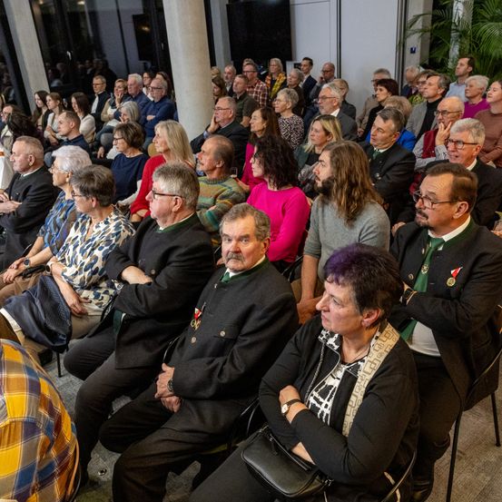 Eine große Gruppe von Menschen ist in einem Raum versammelt, möglicherweise für eine Veranstaltung. Einige tragen Brillen und andere Krawatten. Sie sitzen auf Stühlen, und einige stehen. An der Seite des Raumes befinden sich Pflanzen.