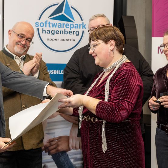 Eine Frau in einem samtroten Kleid erhält ein Dokument von einem Mann im Anzug, während andere klatschen. Sie stehen vor einem Banner mit dem Logo von Softwarepark Hagenberg.