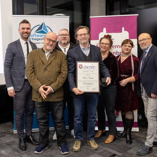 Eine Gruppe von Menschen, darunter Männer und Frauen, steht vor einem Banner. Ein Mann hält einen gerahmten Preis und eine Trophäe.