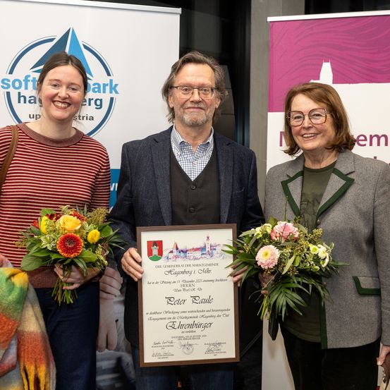 Drei Personen stehen zusammen und halten Blumen und ein gerahmtes Zertifikat. Der Mann in der Mitte hält das Zertifikat und lächelt. Die Frau links und die Frau rechts lächeln ebenfalls und schauen zur Kamera. Hinter ihnen befindet sich ein weißes Banner mit einem Logo.