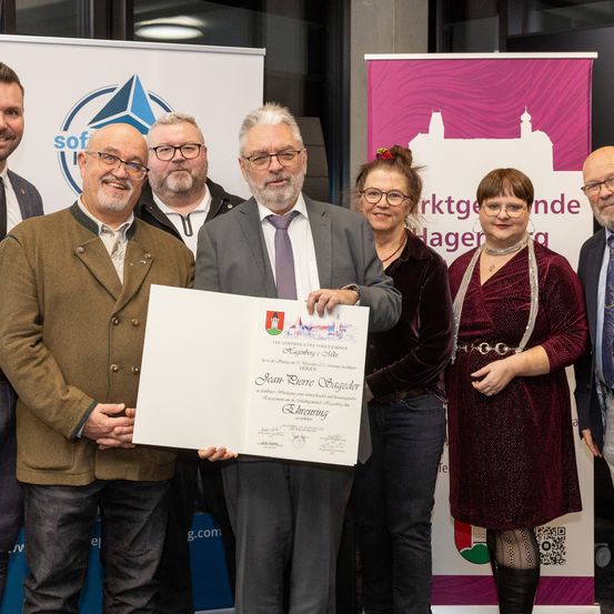 Eine Gruppe von Menschen steht in einem Raum mit einer Bühne, wahrscheinlich für eine Preisverleihung. Der Mann in der Mitte hält ein Zertifikat. Hinter ihm befinden sich zwei Banner, eines mit einem Logo und Text.