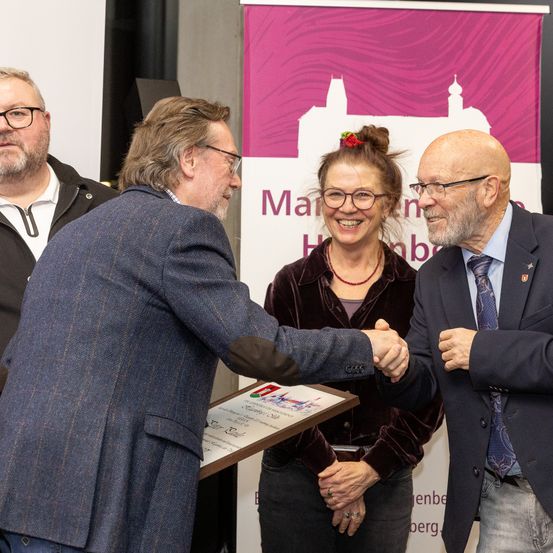 Eine Gruppe von Menschen steht in der Nähe eines Banners, zwei Männer schütteln sich die Hände und eine Frau hält eine Auszeichnung.
