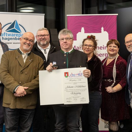 Eine Gruppe von Personen, darunter eine, die ein Zertifikat und eine Medaille hält, lächelt und posiert für ein Foto. Dahinter werden Banner ausgestellt.