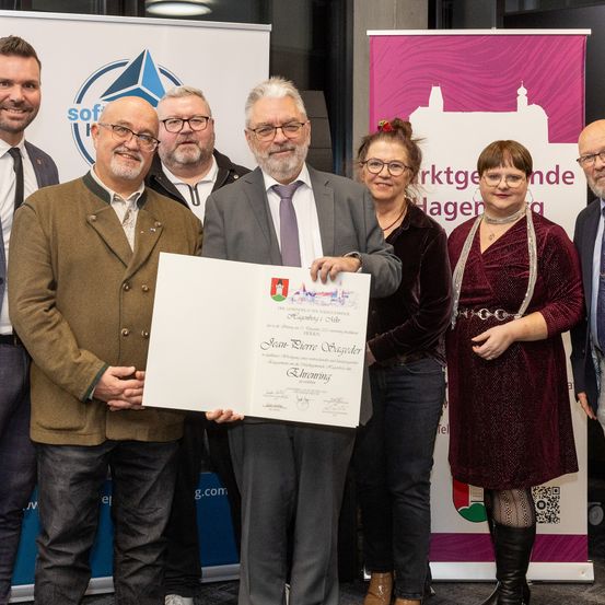 Eine Gruppe von Menschen steht im Innenraum und hält ein Zertifikat. Der Mann in der Mitte präsentiert es einer Frau in einem burgunderroten Kleid. Sie lächeln und posieren für ein Foto.