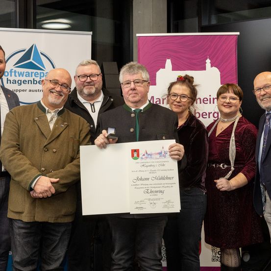 Eine Gruppe von Menschen steht zusammen und hält ein Zertifikat. Der Mann in der Mitte trägt eine Brille und eine Medaille. Der Mann auf der linken Seite trägt einen Mantel. Hinter ihnen ist ein Banner angebracht.