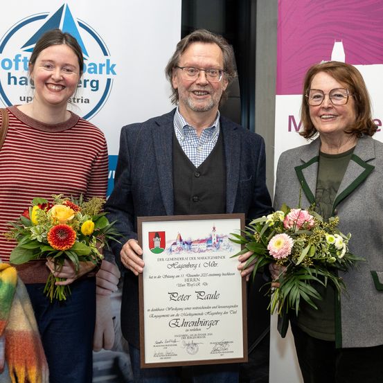 Drei Personen posieren für ein Foto mit einem Zertifikat, alle lächeln. Der Mann hält das Zertifikat mit dem Namen Peter Paule. Die Frauen halten Blumensträuße. Der Hintergrund zeigt ein Logo und Text.