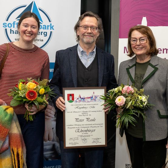 Drei Personen stehen zusammen und halten Blumen, eine in der Mitte hält ein gerahmtes Zertifikat. Der Hintergrund zeigt ein Banner mit Logos und Text, was auf eine Preisverleihung oder Anerkennungszeremonie hindeutet.