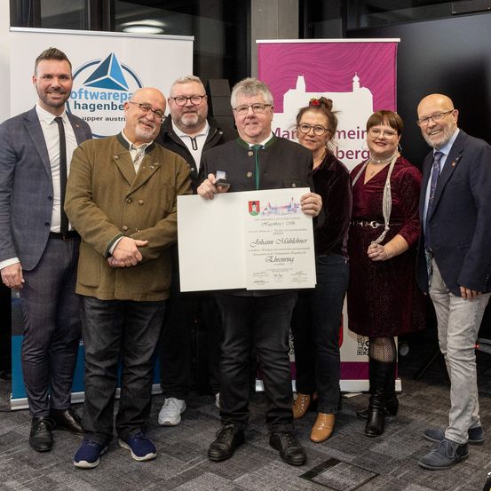 Eine Gruppe von Menschen, darunter zwei Frauen und vier Männer, posiert für ein Foto und hält eine Urkunde in den Händen. Der Mann in der Mitte trägt eine Medaille. Sie stehen vor Bannern mit Logos und Text.