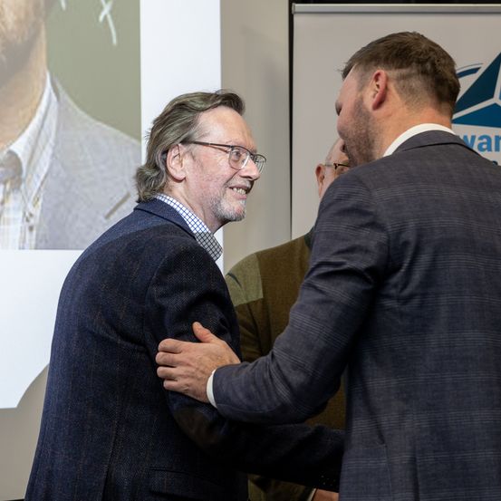 Zwei Männer in Anzügen unterhalten sich. Der Mann auf der linken Seite trägt eine Brille und einen Bart, während der Mann auf der rechten Seite einen Glatzenkopf hat. Sie lächeln und scheinen das Gespräch zu genießen. Hinter ihnen befindet sich ein großes Poster eines Mannes.