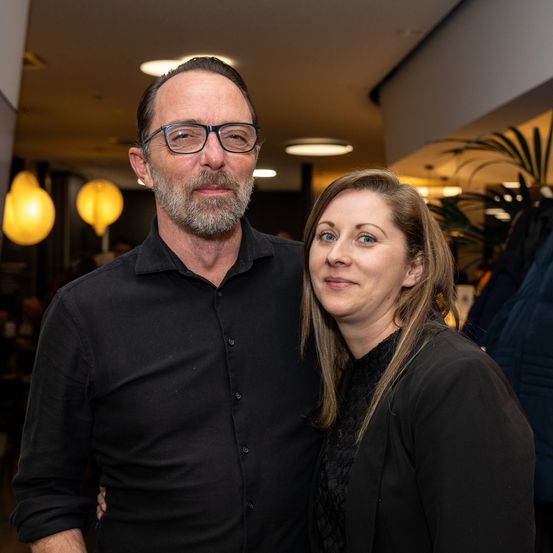 Ein Mann und eine Frau, beide in Schwarz gekleidet, stehen eng beieinander und posieren für ein Foto. Der Mann hat einen Bart und trägt eine Brille. Die Frau hat langes Haar. Hinter ihnen befinden sich Lampen und Pflanzen.