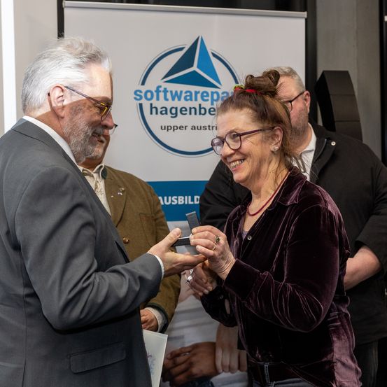 Ein Mann in einem Anzug überreicht eine Auszeichnung an eine lächelnde Frau, die eine Brille und ein lilafarbenes Oberteil trägt. Hinter ihnen steht ein Banner mit der Aufschrift 'softwarepartner hagenbe upper austria'. Ein weiterer Mann in einem schwarzen Jackett beobachtet sie.