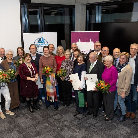 Eine Gruppe von Menschen steht vor einem Banner, wahrscheinlich bei einer Veranstaltung. Einige von ihnen halten Blumen und andere Papiere. Sie alle lächeln und schauen in die Kamera. Hinter ihnen ist eine Glaswand mit einem schwarzen Bildschirm.