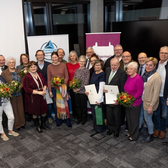 Eine Gruppe von Menschen, wahrscheinlich Mitarbeiter, steht in einem Raum, hält Urkunden und Blumen. Sie scheinen eine Errungenschaft zu feiern.