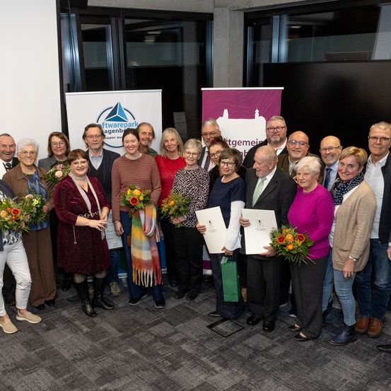 Eine Gruppe von Menschen steht zusammen und hält Urkunden und Blumen. Im Hintergrund sind zwei Banner zu sehen. Sie scheinen eine Leistung zu feiern.