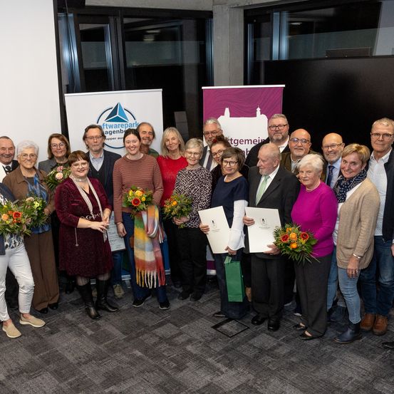 Eine Gruppe von Menschen steht drinnen zusammen, hält Urkunden und Blumensträuße. Einige tragen Brillen, andere Schals. Banner mit Logos stehen hinter ihnen.