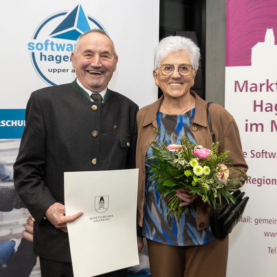 Ein älteres Paar steht vor einem weißen Hintergrund mit einem Logo, lächelt und posiert für ein Foto. Der Mann hält eine Urkunde, die Frau einen Blumenstrauß. Hinter ihnen ist ein rosafarbener Plakat mit dem Text 'Markt im Hagel' zu sehen.