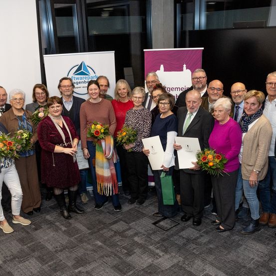Eine Gruppe von Menschen steht drinnen, hält Urkunden und Blumen und ist wahrscheinlich bei einer Preisverleihung. Hinter ihnen sind Banner mit Logos und Text.