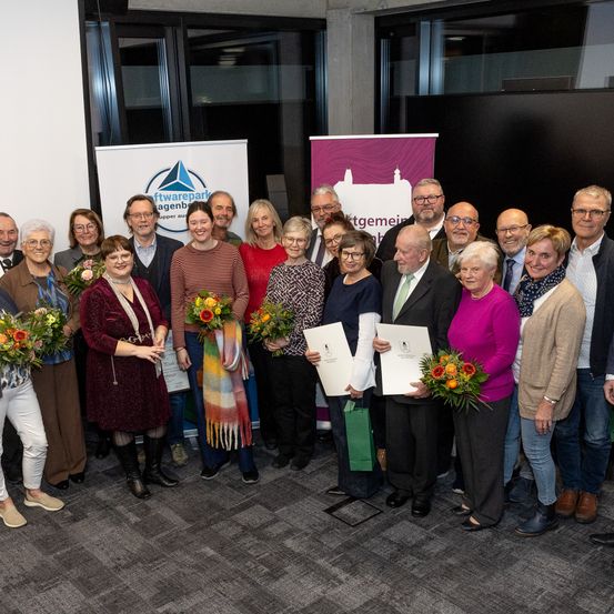 Gruppe von formell gekleideten Personen, die Papiere und Blumen halten, in einem Raum mit Bannern im Hintergrund.