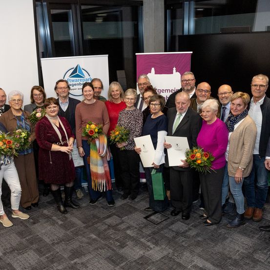 Eine Gruppe von Erwachsenen steht in einem Raum, hält Urkunden und Blumen und lächelt für ein Foto. Banner sind im Hintergrund zu sehen, und einige Personen tragen Brillen.
