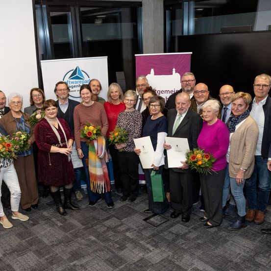 Eine Gruppe von Personen, sowohl Männer als auch Frauen, versammelt sich für ein Foto. Einige halten Urkunden und Blumensträuße. Sie befinden sich in einem Raum mit Bannern und Glasfenstern.