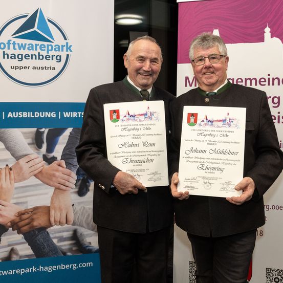 Zwei Männer stehen vor einem Softwarepark Hagenberg Banner, lächelnd und halten Urkunden in den Händen. Ein Mann hat eine Urkunde mit dem Titel 'Hubert Ronn' und der andere hat eine Urkunde mit dem Titel 'Johann Mühlhehner'. Sie sind in schwarzen Anzügen mit grünen Krawatten gekleidet.