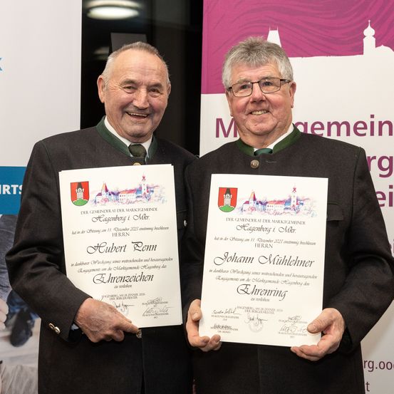 Zwei ältere Männer lächeln und halten Urkunden vor einem Banner. Eine liest 'Hubert Penn' und die andere 'Johann Mühlhehner'. Beide tragen traditionelle Kleidung. Hinter ihnen ein teilweise sichtbares Banner mit einer Burg und Text.