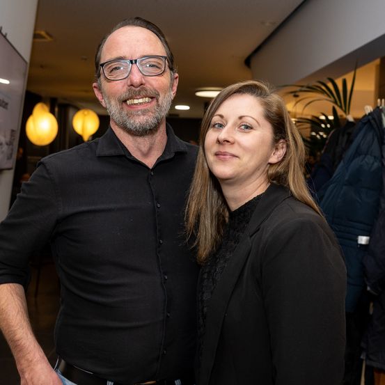 Ein Mann und eine Frau stehen eng nebeneinander in einem Raum und lächeln für ein Foto. Der Mann trägt eine Brille und ein schwarzes Hemd, während die Frau lange Haare und eine schwarze Jacke hat. Hinter ihnen steht eine Pflanze und Jacken hängen an einem Ständer.