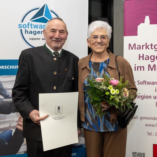 Ein älterer Mann und eine ältere Frau stehen vor einem Banner, lächeln und posieren für ein Foto. Der Mann hält eine Urkunde und die Frau einen Blumenstrauß. Hinter ihnen steht ein Banner mit der Aufschrift 'Marktgemeinde Hagenberg'.