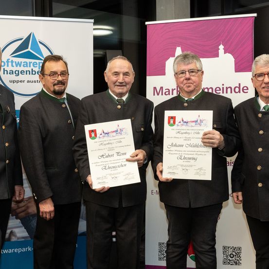 Fünf Männer in traditioneller Kleidung stehen vor einem rosafarbenen Banner. Zwei von ihnen halten Urkunden in den Händen, und sie alle lächeln für ein Foto. Das Banner hinter ihnen hat ein Stadtwappen und das Wort 'Marktgemeinde'.