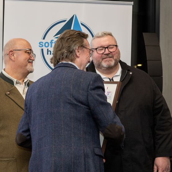 Drei Männer stehen vor einem Banner mit Logo. Der Mann in der Mitte hält ein gerahmtes Bild und scheint einen Preis zu vergeben.