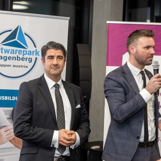 Zwei Männer in Anzügen und Krawatten stehen vor einem Mikrofon. Ein Mann hält das Mikrofon. Hinter ihnen sind Banner mit Logos und Texten angebracht.