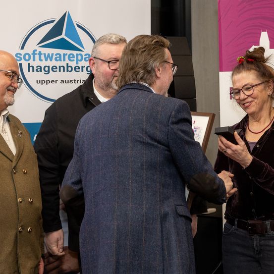 Vier Personen stehen vor einem Hintergrund mit dem Logo von Software Hagenberg. Ein Mann schüttelt einer Frau, die eine Auszeichnung hält, die Hand.