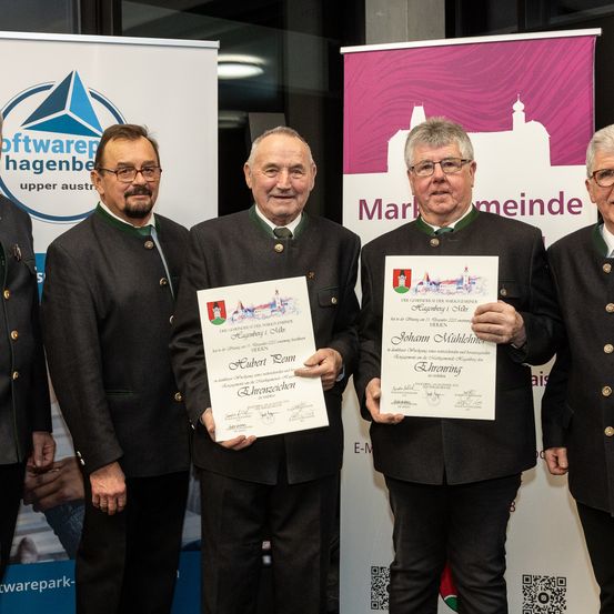 Fünf Männer in formeller Kleidung stehen hinter einem Banner und halten Urkunden. Der Mann auf der rechten Seite hält eine Urkunde für Johann Mühlmeier, datiert auf Dezember 2022. Zwei Männer auf der linken Seite halten Urkunden für Hubert Denn und Ehrenzeichen.