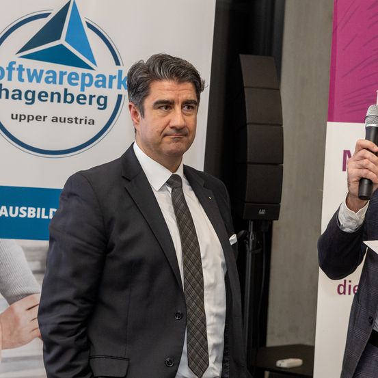 Ein Mann in Anzug und Krawatte steht vor einem Banner mit einem Mikrofon auf der rechten Seite. Hinter ihm steht ein Lautsprecher und eine Person, die Hände schüttelt.