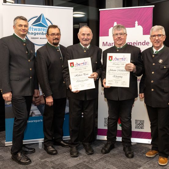 Fünf Männer in traditioneller österreichischer Kleidung stehen zusammen und halten Urkunden. Sie stehen vor Bannern mit Logos und Text. Ein Mann hält eine Urkunde mit dem Titel 'Johann Mittelstein'. Ein anderer Mann hält eine Urkunde mit dem Titel 'Eduard'. Sie tragen alle Brillen und Schuhe.
