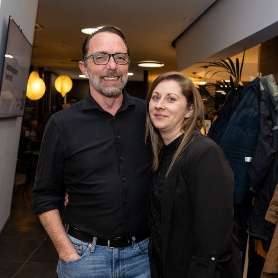 Ein Mann und eine Frau stehen nah beieinander und lächeln, mit einem Geschäft im Hintergrund, das einen Kleiderständer mit Jacken und ein Schild an der Wand zeigt.