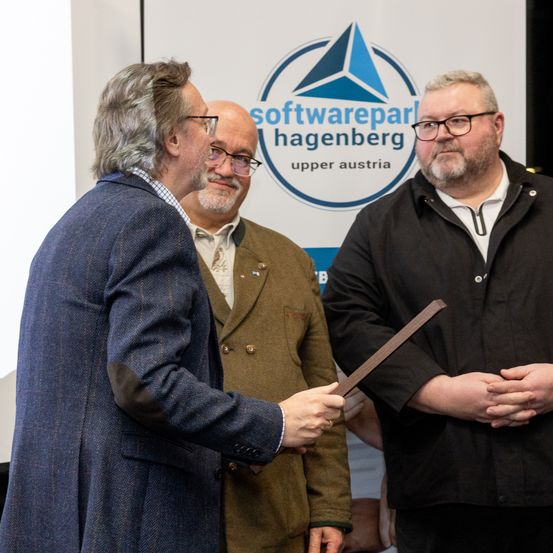 Drei Männer stehen vor einem Banner mit Logo. Der Mann in der Mitte hält einen Holzstab.