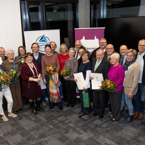 Eine Gruppe von Erwachsenen, einige halten Blumen und Urkunden, stehen zusammen in einem Raum, wahrscheinlich bei einer Preisverleihung. Sie stehen vor Bannern mit Logos.